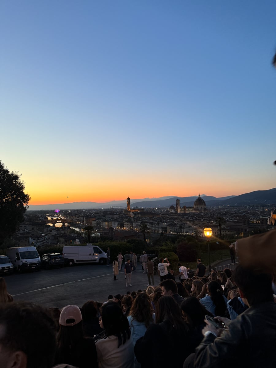A crowd of people sits on steps overlooking Florence at sunset, enjoying the cityscape-including the Duomo and Palazzo Vecchio-against a colorful sky of blue and orange hues, capturing the essence of exploring Florence in One Day.