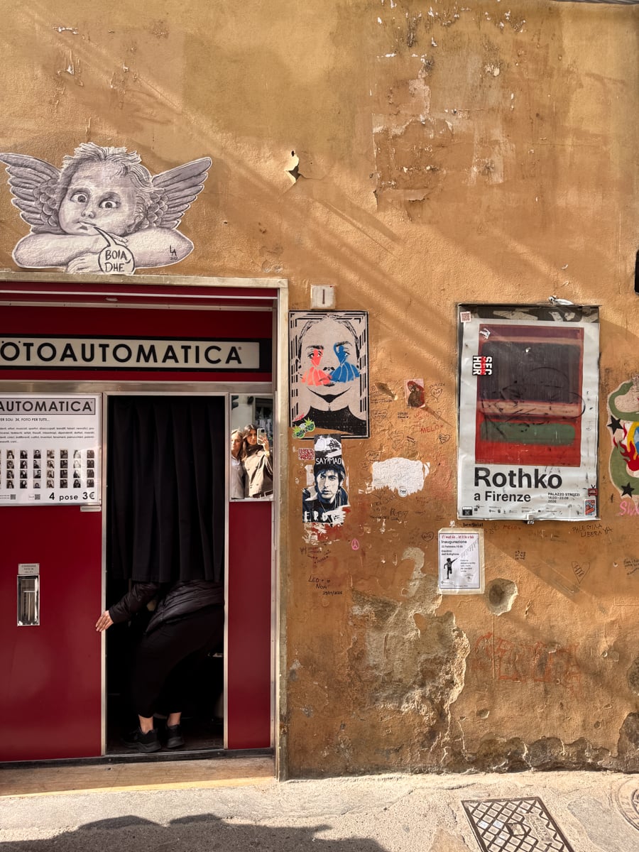 A person partially enters a vintage red photo booth set against a weathered, graffiti-covered wall with posters—including one for a Rothko exhibit—reminiscent of the vibrant atmosphere found while exploring Florence in One Day.