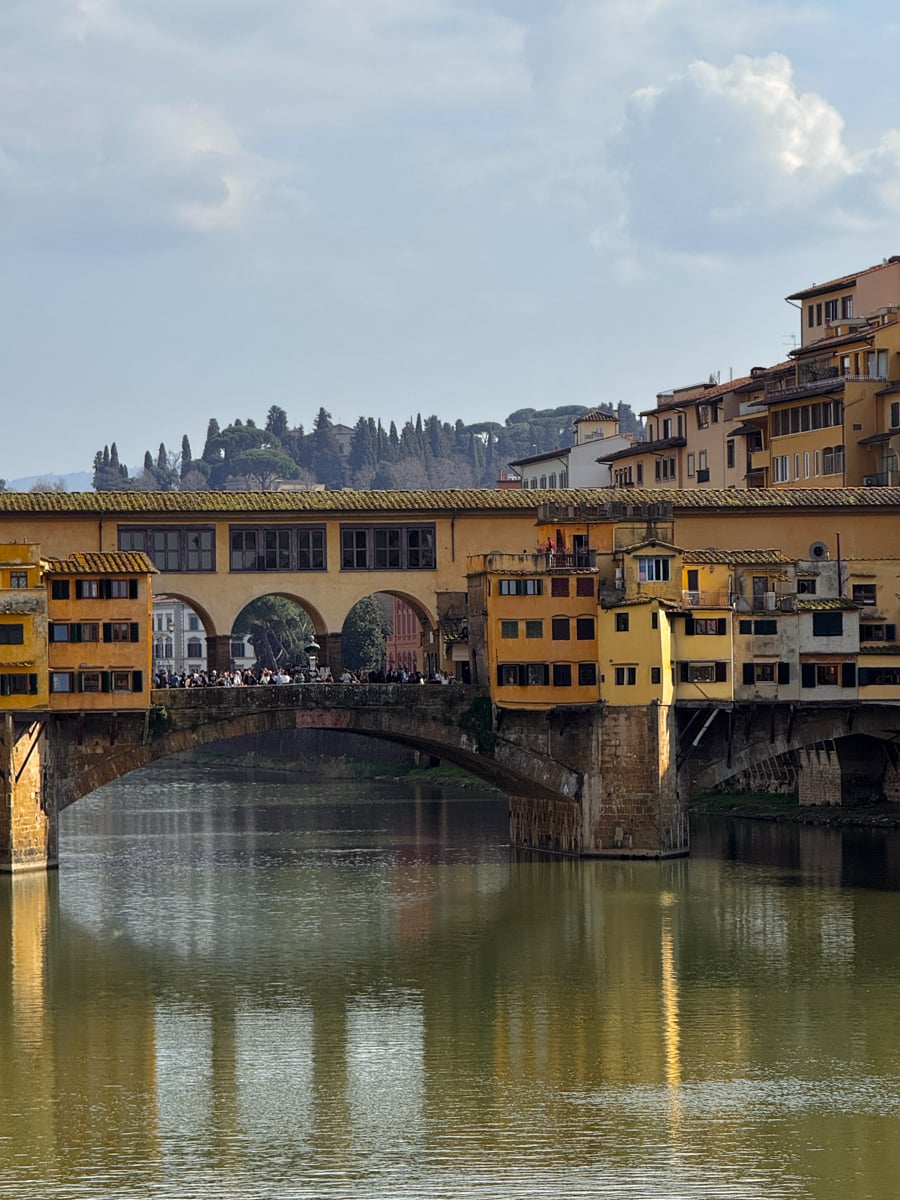 A historic stone bridge with shops along its span crosses a calm river in Florence in One Day, surrounded by yellow buildings. Trees and hills are visible in the background under a cloudy sky.