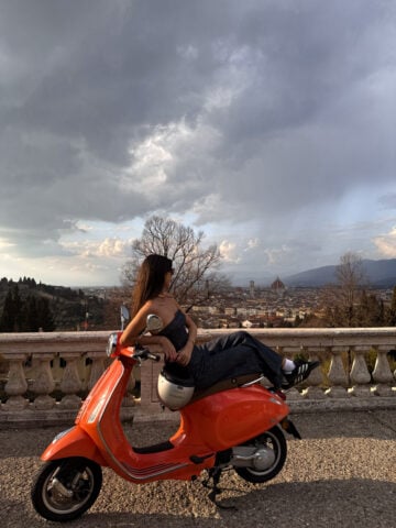A woman sits sideways on an orange scooter, gazing at a scenic city view with distant mountains and dramatic clouds—a perfect snapshot of Florence in One Day, blending urban landscape with natural beauty.