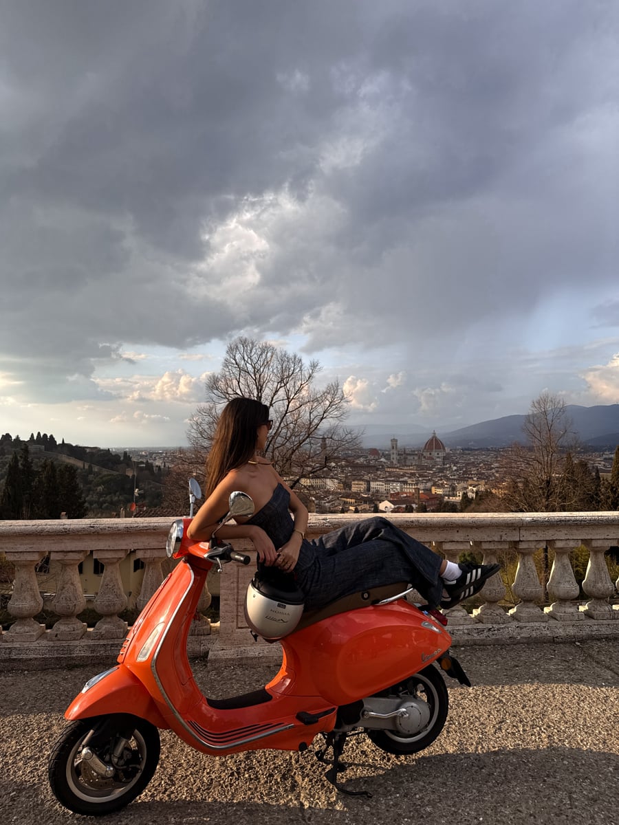 A woman sits sideways on an orange scooter, gazing at a scenic city view with distant mountains and dramatic clouds—a perfect snapshot of Florence in One Day, blending urban landscape with natural beauty.