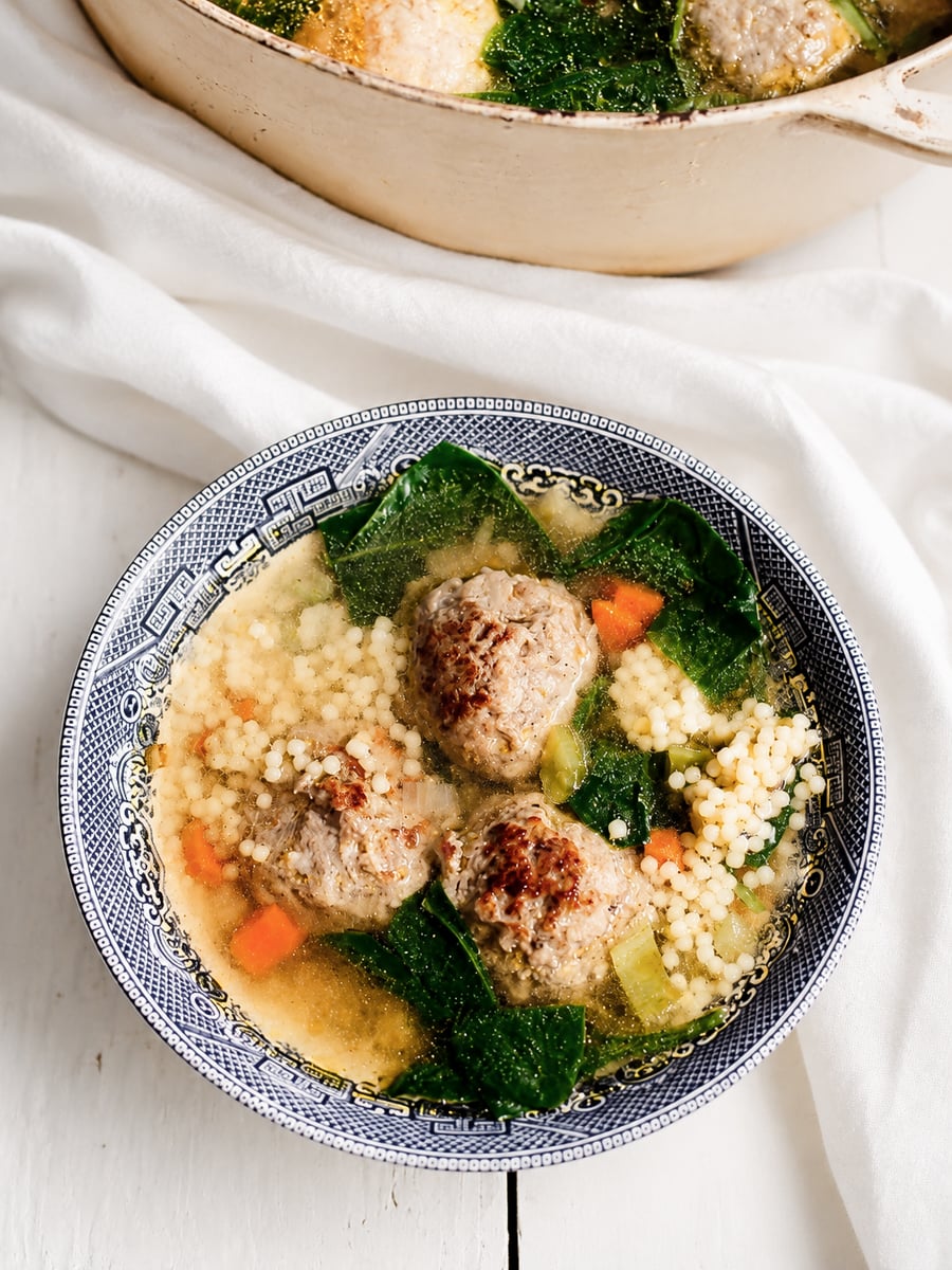 Blue-patterned bowl of Italian Wedding Soup with meatballs, small round pasta, carrots, celery, and dark leafy greens in broth, placed on a white surface with a white cloth and a pot of soup in the background.