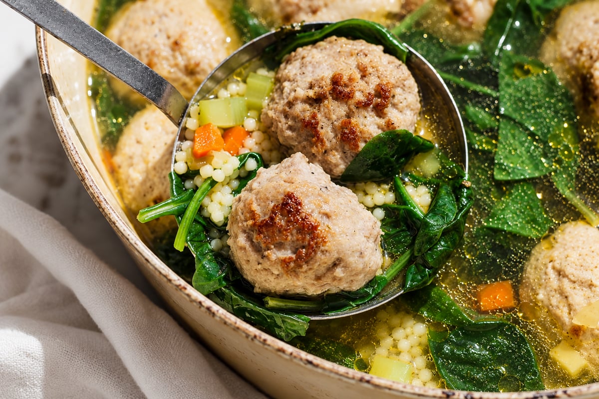 A ladle lifts meatballs, spinach, carrots, celery, and tiny pasta from a pot of Italian Wedding Soup. The broth is clear and golden, with visible vegetables and greens.