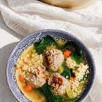 A bowl of Italian Wedding Soup, featuring savory meatballs, couscous, carrots, leafy greens, and flavorful broth, served in a blue-patterned bowl on a white wooden surface.