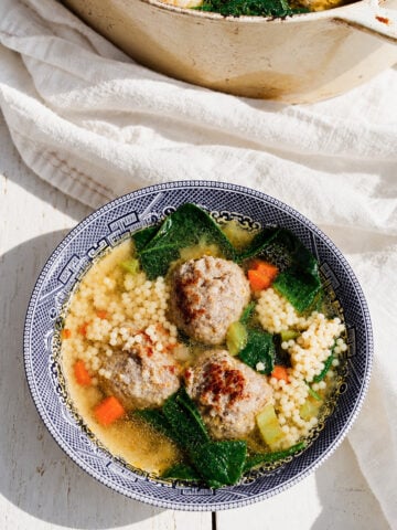 A bowl of Italian Wedding Soup, featuring savory meatballs, couscous, carrots, leafy greens, and flavorful broth, served in a blue-patterned bowl on a white wooden surface.