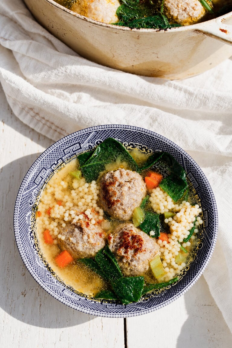 A bowl of Italian Wedding Soup, featuring savory meatballs, couscous, carrots, leafy greens, and flavorful broth, served in a blue-patterned bowl on a white wooden surface.