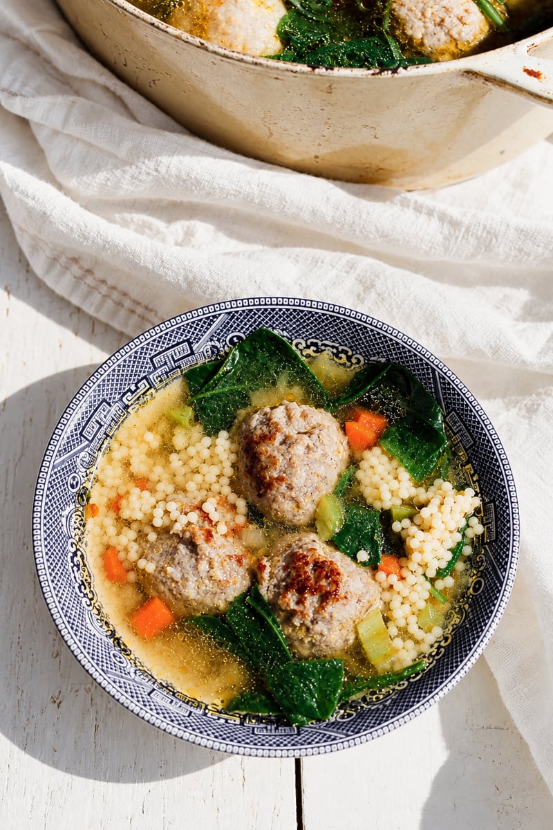 A bowl of Italian Wedding Soup, featuring savory meatballs, couscous, carrots, leafy greens, and flavorful broth, served in a blue-patterned bowl on a white wooden surface.