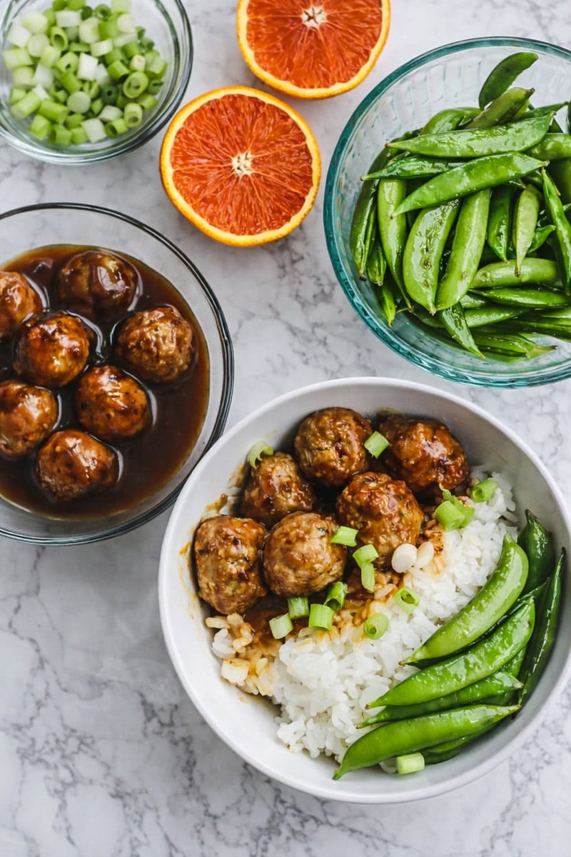 A bowl of white rice is topped with Orange Chicken Meatballs, sliced green onions, and snap peas. Surrounding the bowl are dishes of saucy meatballs, crisp snap peas, chopped green onions, and vibrant halved blood oranges.