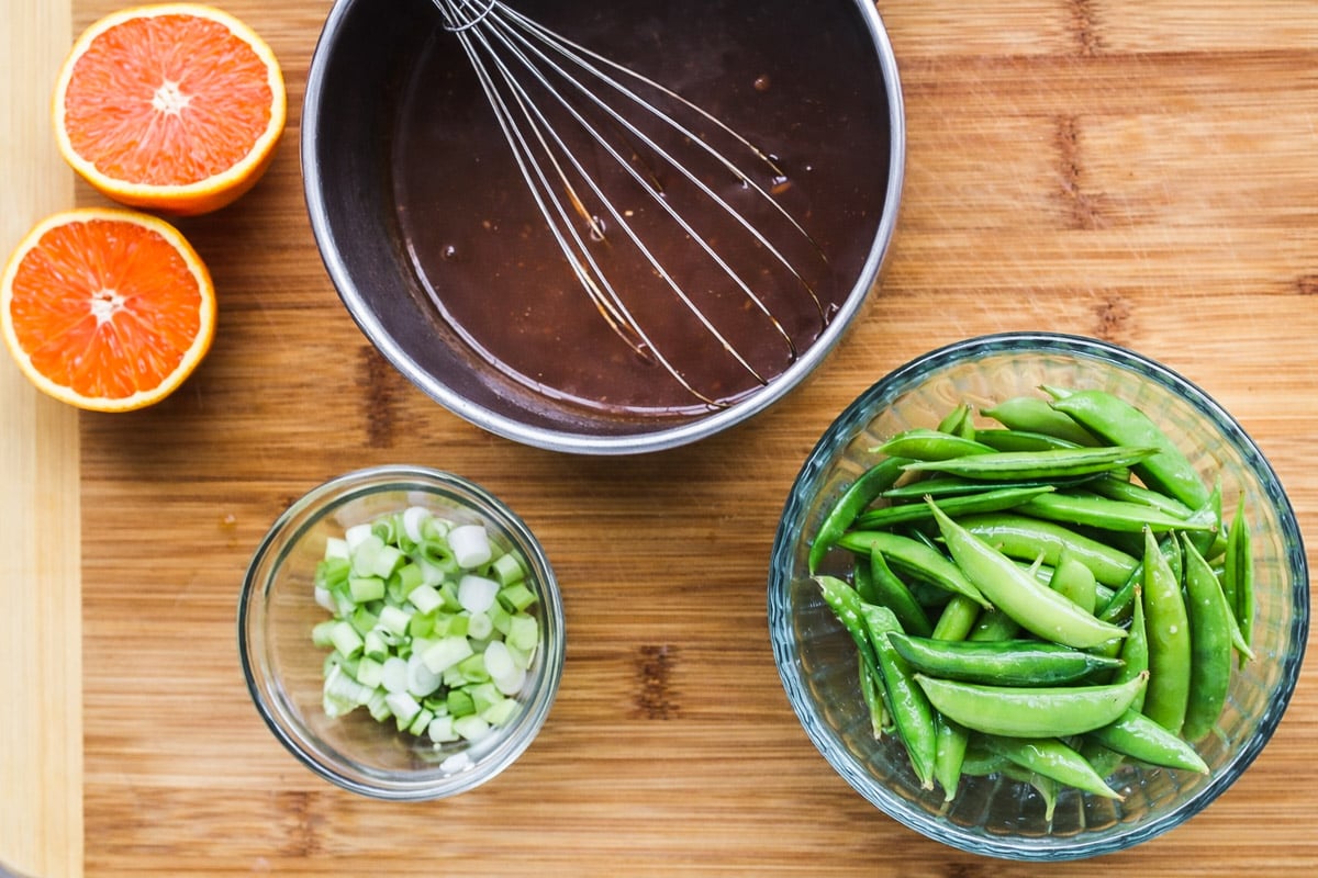 A cutting board with halved oranges, a bowl of sugar snap peas, chopped green onions in a small bowl, and a mixing bowl with dark sauce and a whisk-perfect ingredients for preparing Orange Chicken Meatballs.