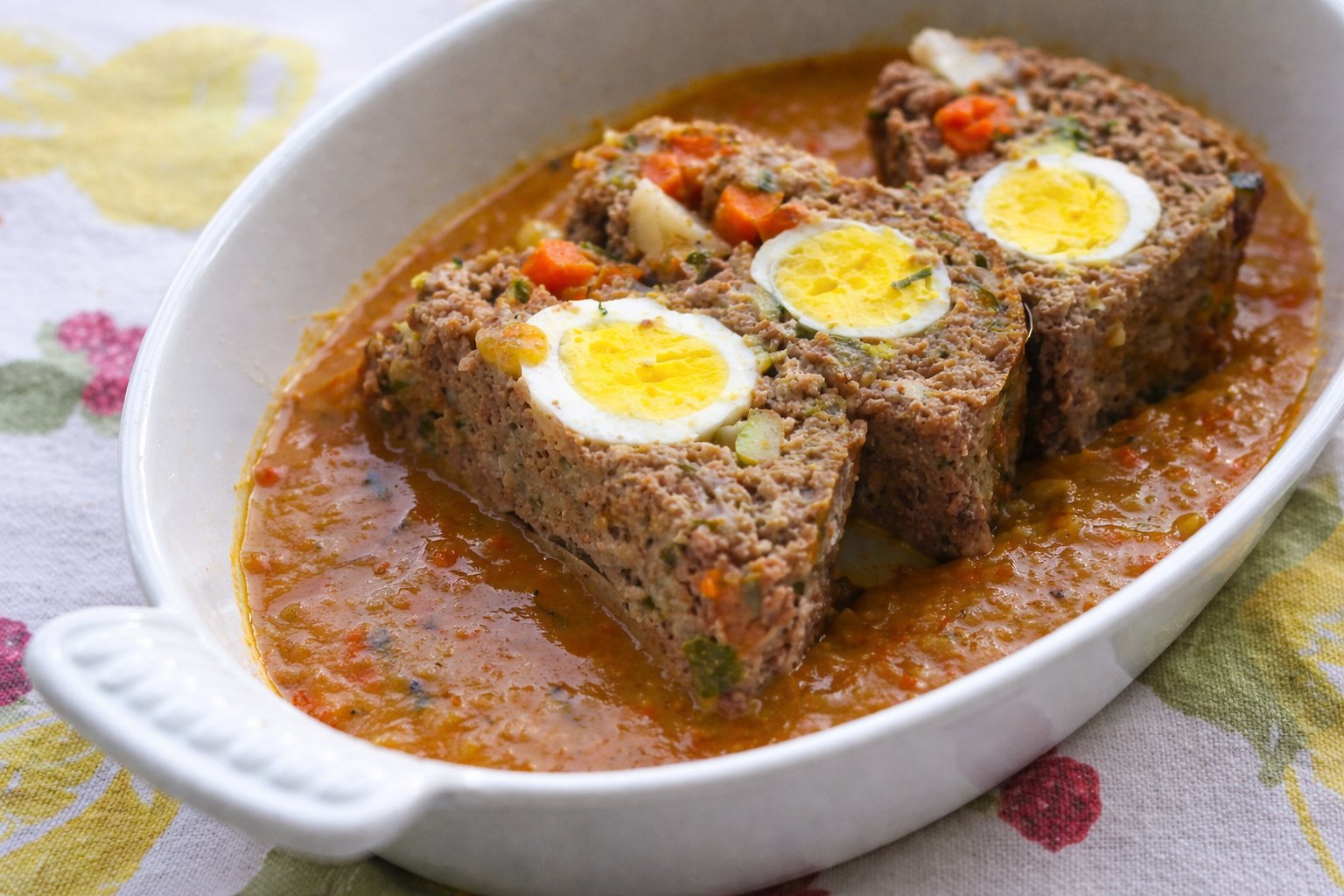 Oval dish with slices of Italian meatloaf containing whole boiled eggs in the center, topped with a chunky tomato-based sauce. The dish is placed on a patterned tablecloth.