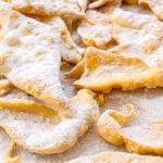 Golden, crispy Chiacchiere are dusted with powdered sugar and arranged in a scattered pile on a white surface.