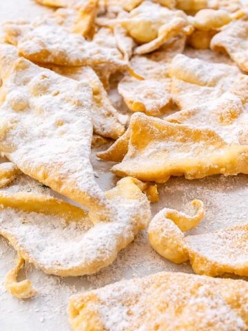 Golden, crispy Chiacchiere are dusted with powdered sugar and arranged in a scattered pile on a white surface.