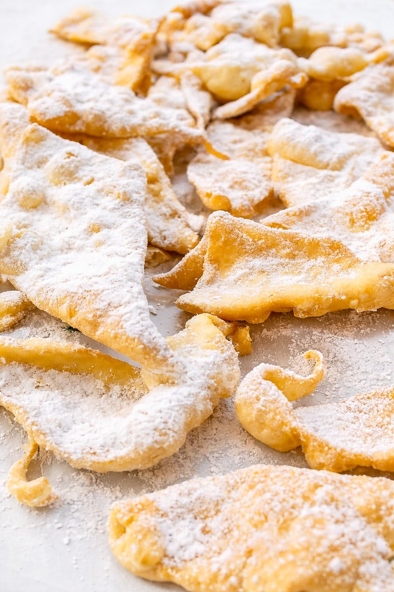 Golden, crispy Chiacchiere are dusted with powdered sugar and arranged in a scattered pile on a white surface.