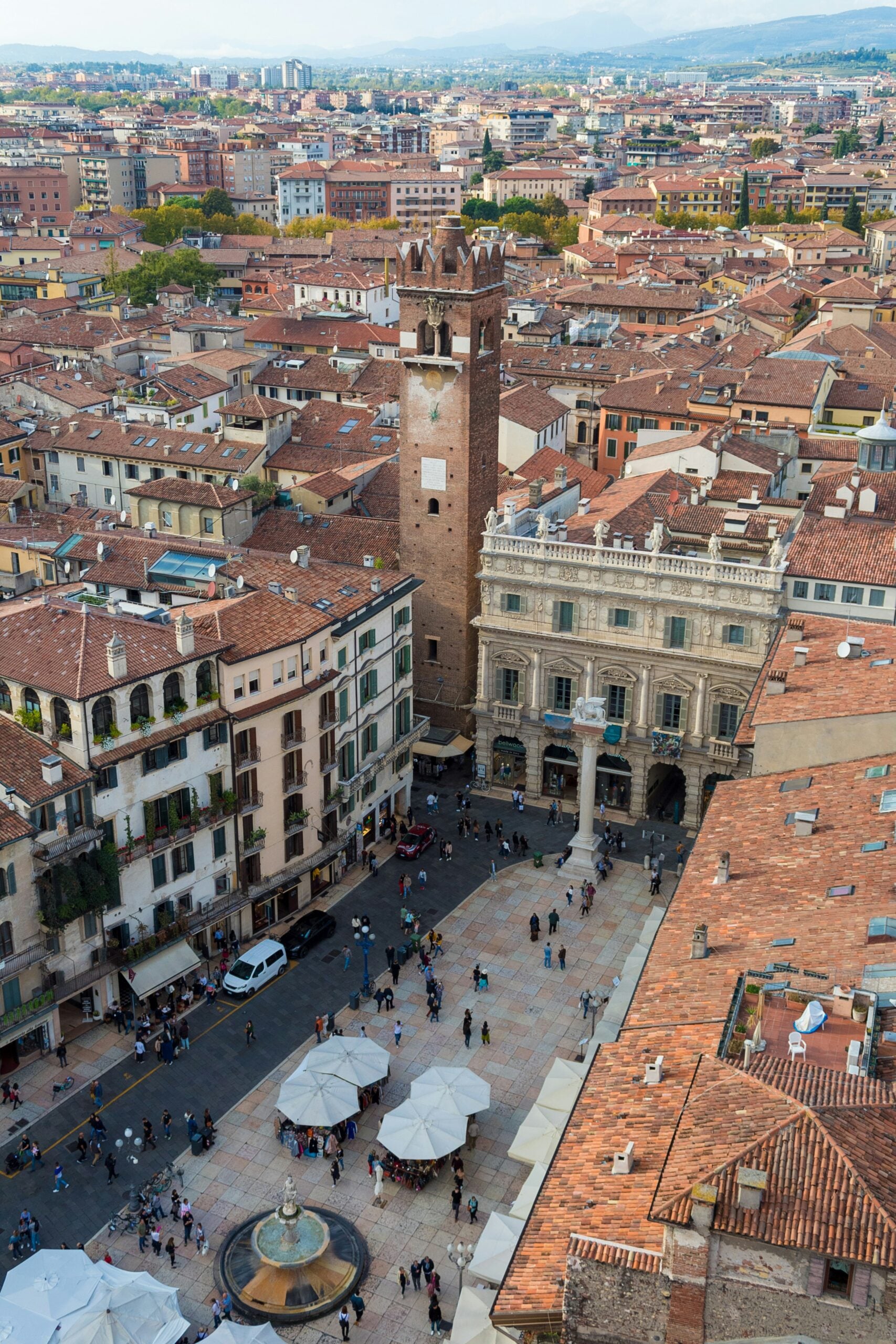 A panoramic aerial shot of Verona, Italy, capturing the bustling Piazza delle Erbe as seen from the top of Torre dei Lamberti. The view showcases the vibrant heart of Verona with its colorful market stalls, historic buildings, and the iconic fountain of Madonna Verona. The surrounding cityscape unfolds with a mix of medieval architecture, red-tiled rooftops, and narrow cobblestone streets. In the distance, the Adige River winds through the city, framed by lush green hills. This image beautifully encapsulates Verona’s blend of history, culture, and natural beauty, making it a captivating destination for travelers.