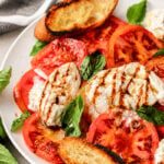 A plate of Caprese salad with fresh tomato slices, mozzarella cheese, basil leaves, a drizzle of balsamic glaze, and toasted bread sits invitingly. A striped napkin is partially visible in the background.