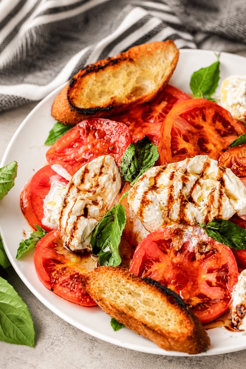 A plate of Caprese salad with fresh tomato slices, mozzarella cheese, basil leaves, a drizzle of balsamic glaze, and toasted bread sits invitingly. A striped napkin is partially visible in the background.
