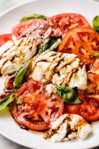 Sliced tomatoes, fresh mozzarella, and basil leaves are arranged on a white plate, then drizzled with balsamic glaze and olive oil for a classic Caprese salad bursting with fresh flavors.