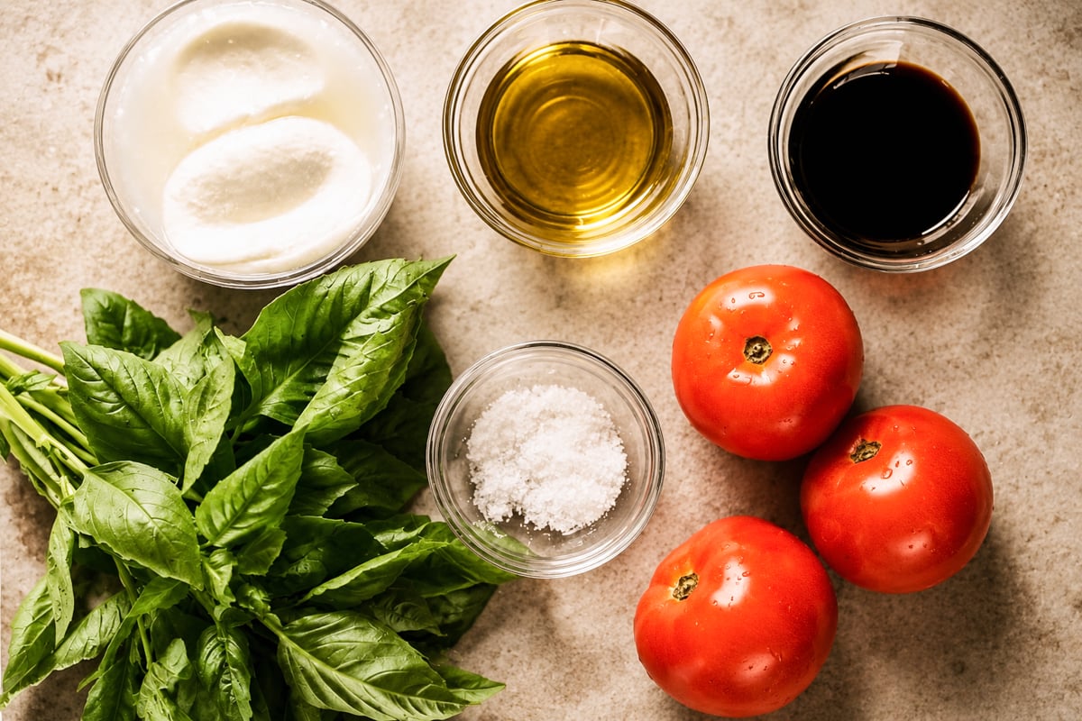 Fresh basil leaves, three whole tomatoes, mozzarella cheese, olive oil, balsamic vinegar, and salt are beautifully arranged on a light countertop—perfect ingredients for a classic Caprese salad.
