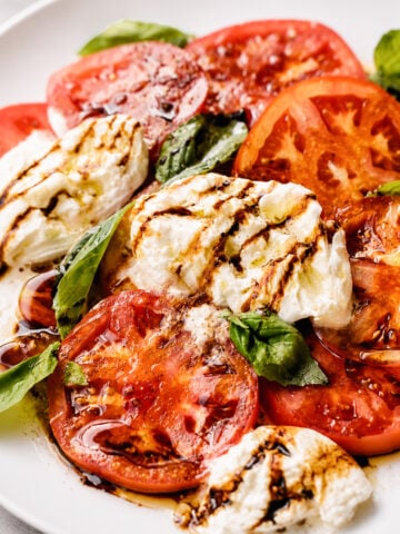 Sliced tomatoes, fresh mozzarella, and basil leaves are arranged on a white plate, then drizzled with balsamic glaze and olive oil for a classic Caprese salad bursting with fresh flavors.