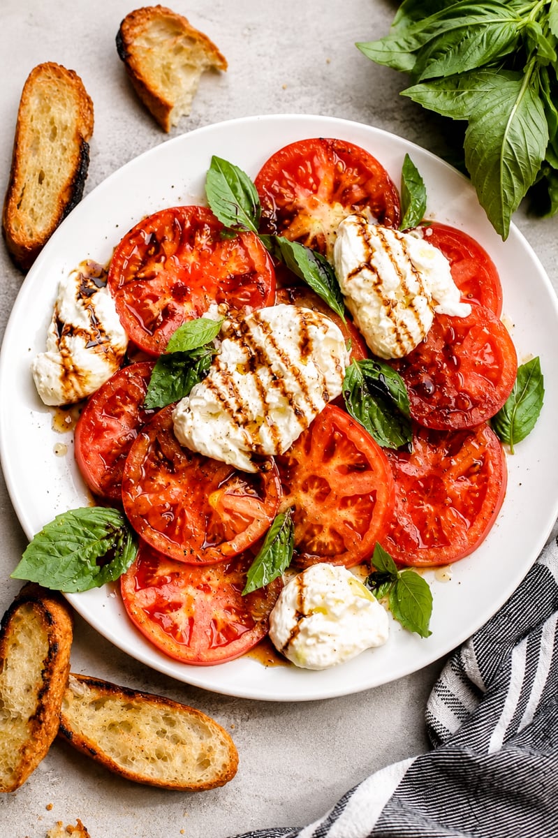 A classic Caprese salad features sliced tomatoes, fresh mozzarella, and basil leaves drizzled with balsamic glaze, surrounded by toasted bread slices and a bunch of basil on the side. A striped cloth napkin is partially visible.