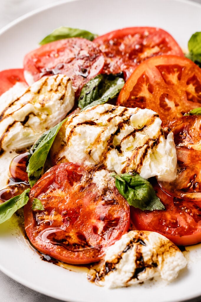 Sliced tomatoes, fresh mozzarella, and basil leaves are arranged on a white plate, then drizzled with balsamic glaze and olive oil for a classic Caprese salad bursting with fresh flavors.