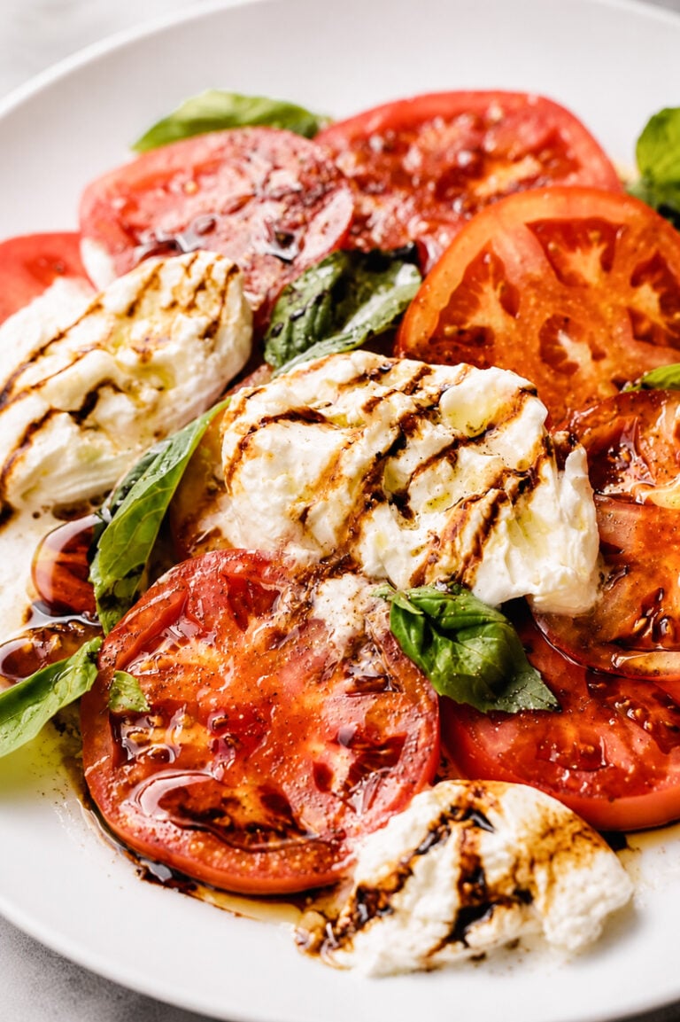 Sliced tomatoes, fresh mozzarella, and basil leaves are arranged on a white plate, then drizzled with balsamic glaze and olive oil for a classic Caprese salad bursting with fresh flavors.