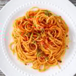 A plate of caramelized shallot pasta topped with tomato sauce and garnished with chopped parsley, served on a white dish with a beaded rim, placed on a rustic wooden table.