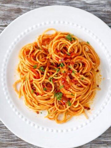 A plate of caramelized shallot pasta topped with tomato sauce and garnished with chopped parsley, served on a white dish with a beaded rim, placed on a rustic wooden table.