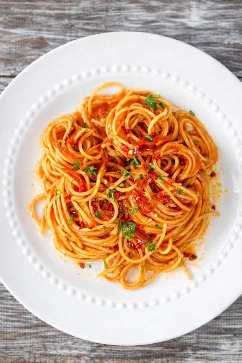 A plate of caramelized shallot pasta topped with tomato sauce and garnished with chopped parsley, served on a white dish with a beaded rim, placed on a rustic wooden table.