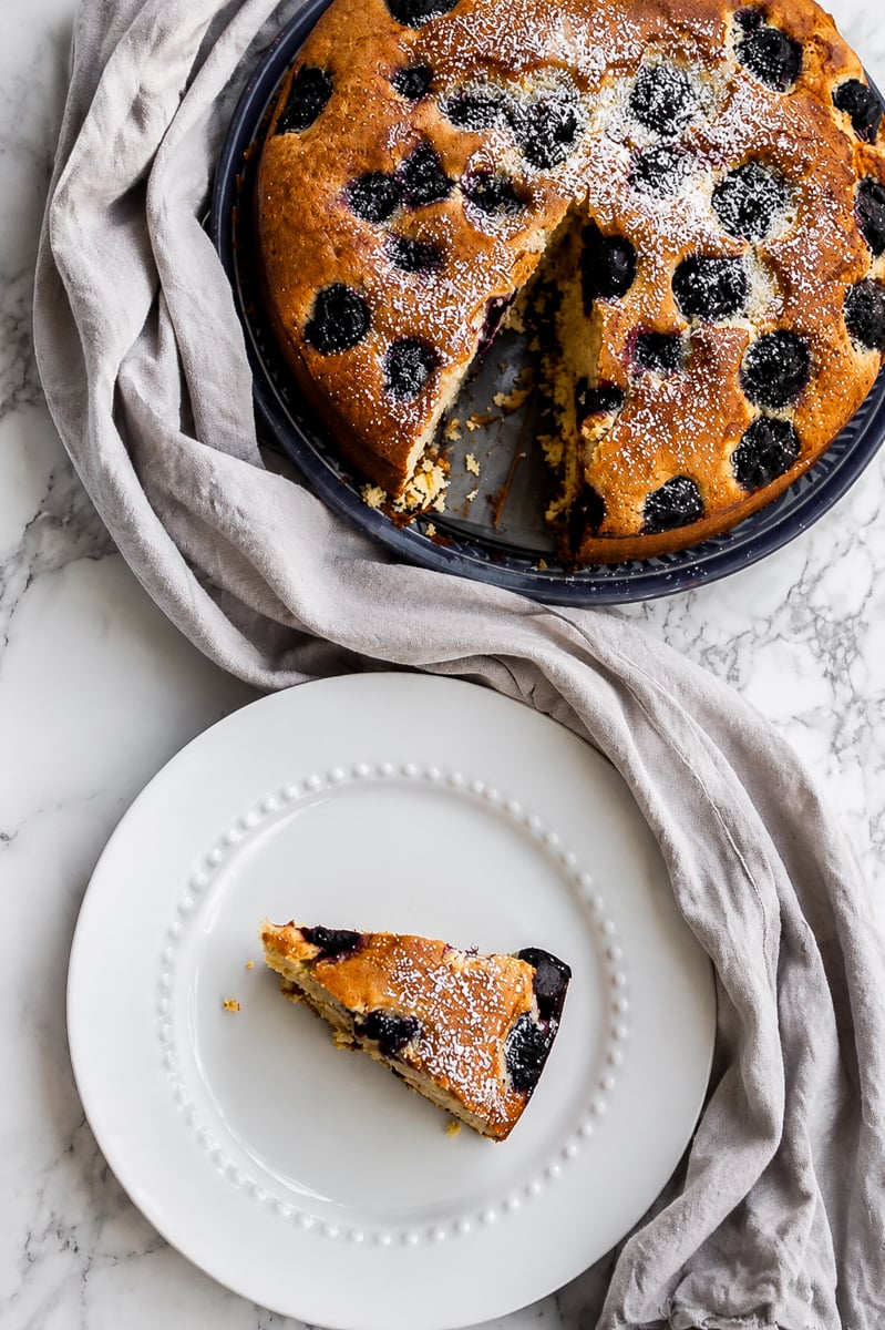 A round cherry cake with a slice missing sits on a tray, topped with powdered sugar. The missing slice is placed on a white plate next to the cake, all arranged on a marble surface with a gray cloth.