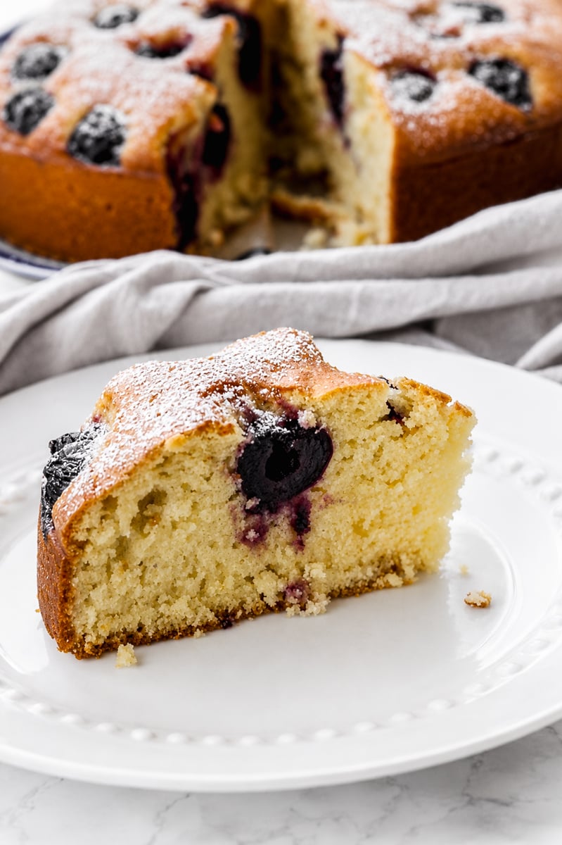 A slice of moist, golden cherry cake with dark berries inside, dusted with powdered sugar, sits on a white plate. In the background, the remaining cake brimming with cherries is visible alongside a light gray cloth.