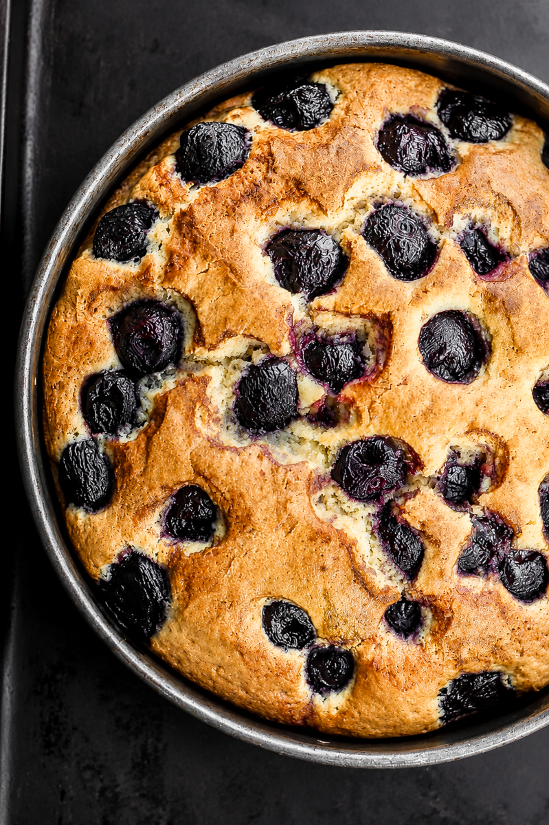 A round, golden-brown Cherry Cake filled with baked dark cherries, shown in a metal cake pan on a dark surface.