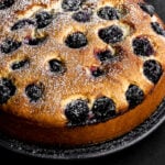 A round cherry cake dusted with powdered sugar sits on a dark plate against a black background. The golden-brown cake is studded with whole blackberries on top.