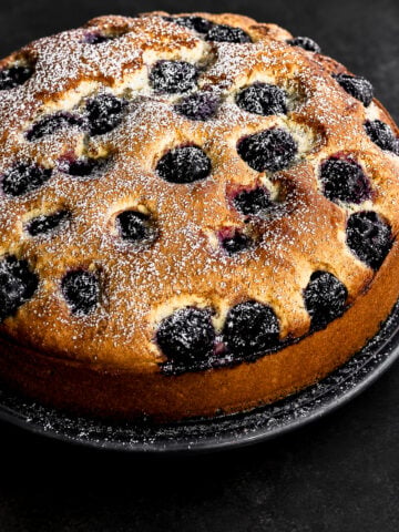 A round cherry cake dusted with powdered sugar sits on a dark plate against a black background. The golden-brown cake is studded with whole blackberries on top.