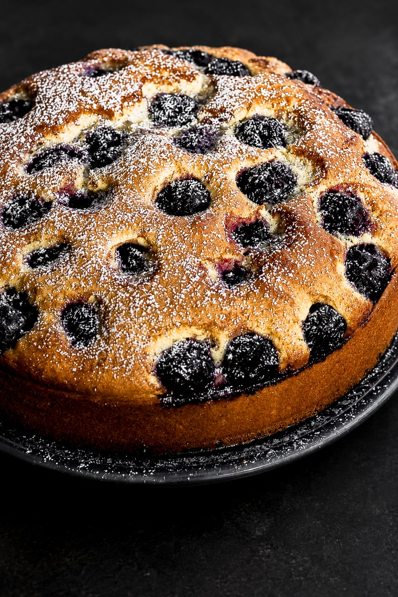 A round cherry cake dusted with powdered sugar sits on a dark plate against a black background. The golden-brown cake is studded with whole blackberries on top.