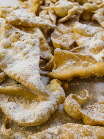 A close-up of several pieces of golden, fried chiacchiere dusted with powdered sugar, overlapping on a flat surface.
