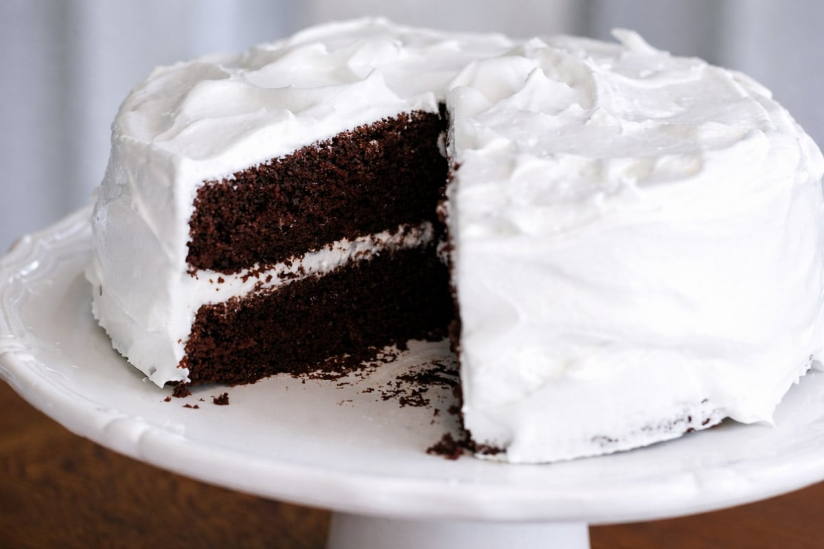 A Chocolate Espresso Layer Cake with two rich layers and white frosting sits on a white cake stand. A large slice has been removed, showing off the dark, espresso-infused cake and creamy frosting between each layer.