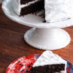 A slice of Chocolate Espresso Layer Cake with white frosting sits on a colorful red and pink plate, while the rest of the cake, with a wedge missing, is displayed on a white cake stand in the background.