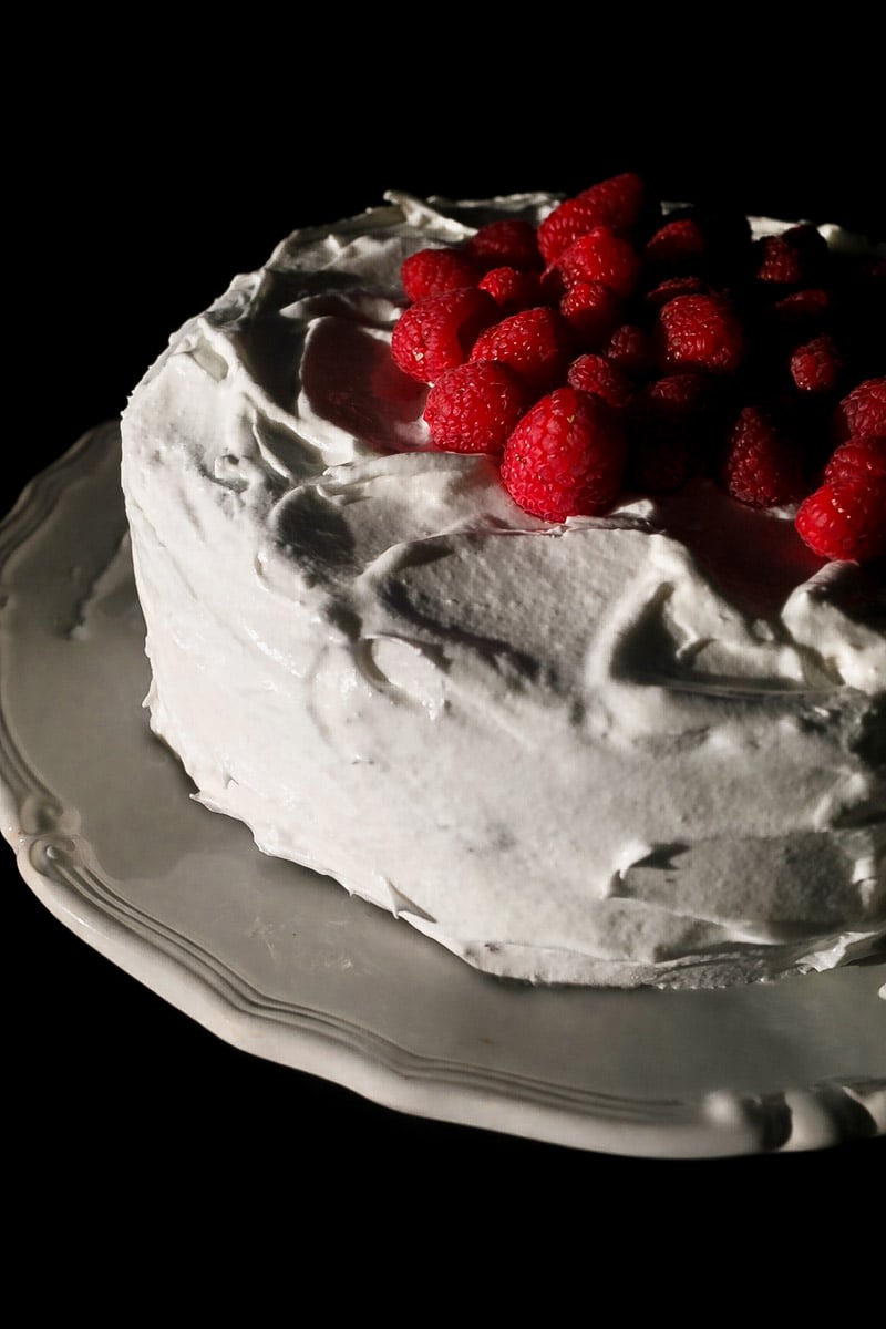 A Chocolate Espresso Layer Cake with white whipped frosting, topped with a cluster of fresh raspberries, is displayed on a decorative white plate against a dark background.