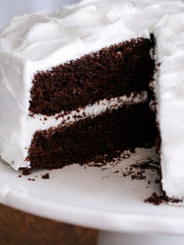 A Chocolate Espresso Layer Cake with two layers and white frosting sits on a white cake stand. A large slice is cut and slightly removed, revealing the cake's moist, rich interior infused with espresso and creamy frosting layers.