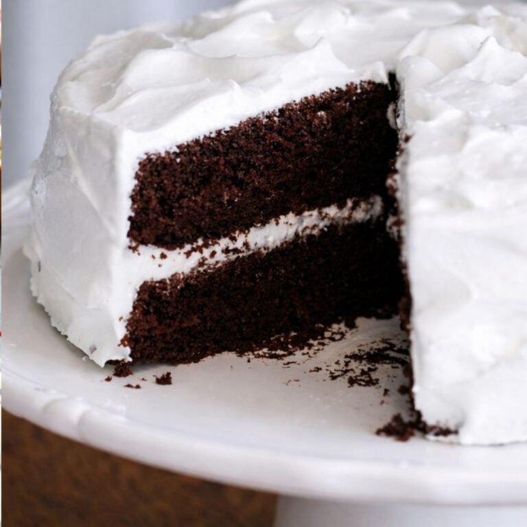A Chocolate Espresso Layer Cake with two layers and white frosting sits on a white cake stand. A large slice is cut and slightly removed, revealing the cake’s moist, rich interior infused with espresso and creamy frosting layers.