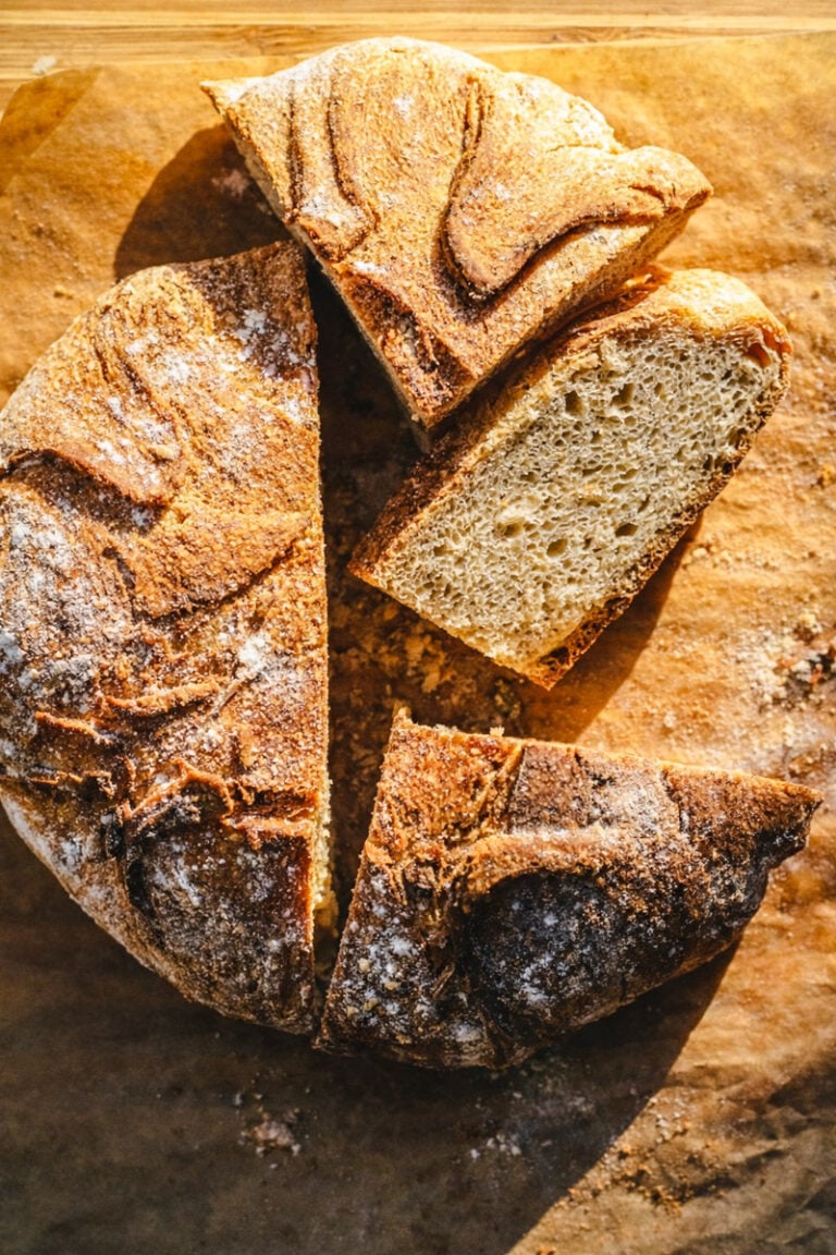 A round loaf of No Knead Italian Bread rests on parchment paper with three slices cut out, revealing a golden, crusty exterior and a soft, airy crumb inside.