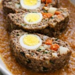 Three slices of Italian meatloaf stuffed with hard-boiled eggs, carrots, and vegetables are served in an oval dish on a bed of tomato-based sauce.