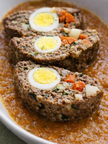 Three slices of Italian meatloaf stuffed with hard-boiled eggs, carrots, and vegetables are served in an oval dish on a bed of tomato-based sauce.