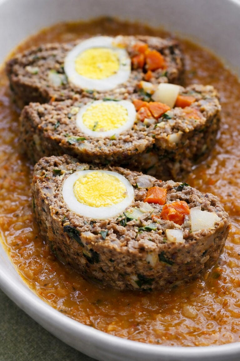 Three slices of Italian meatloaf stuffed with hard-boiled eggs, carrots, and vegetables are served in an oval dish on a bed of tomato-based sauce.