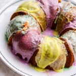 A King Cake with colorful purple, yellow, and green icing sits on a white plate. A small plastic baby figurine is placed on top of the cake.