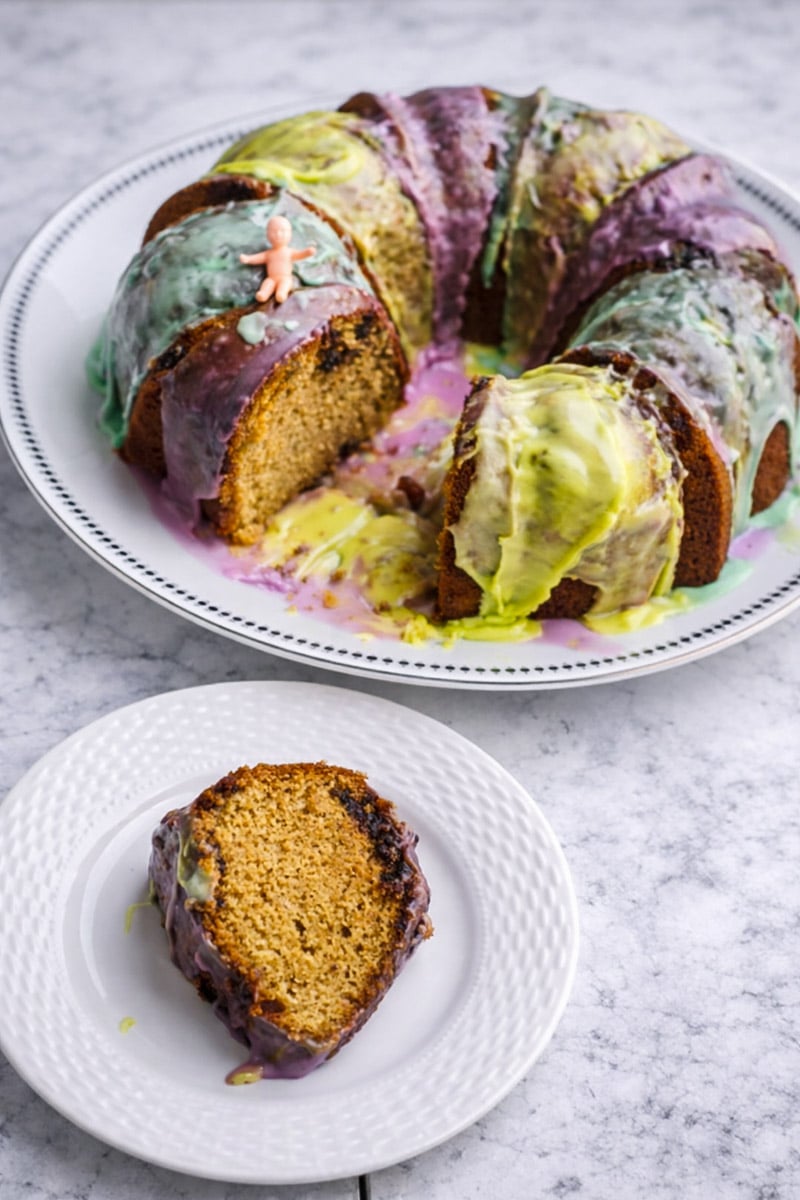 A sliced King Cake with colorful icing and a small plastic baby on top sits on a white plate. A single slice of King Cake is served on a separate small white plate in front of the cake.