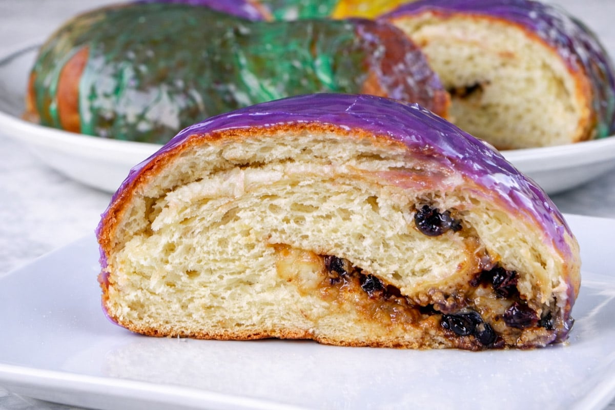 A slice of King Cake with a flaky, golden interior, filled with dark raisins or berries and topped with purple icing, sits on a white plate. More colorful King Cake slices are visible in the background.