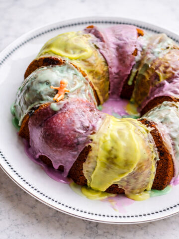 A King Cake with colorful purple, yellow, and green icing sits on a white plate. A small plastic baby figurine is placed on top of the cake.
