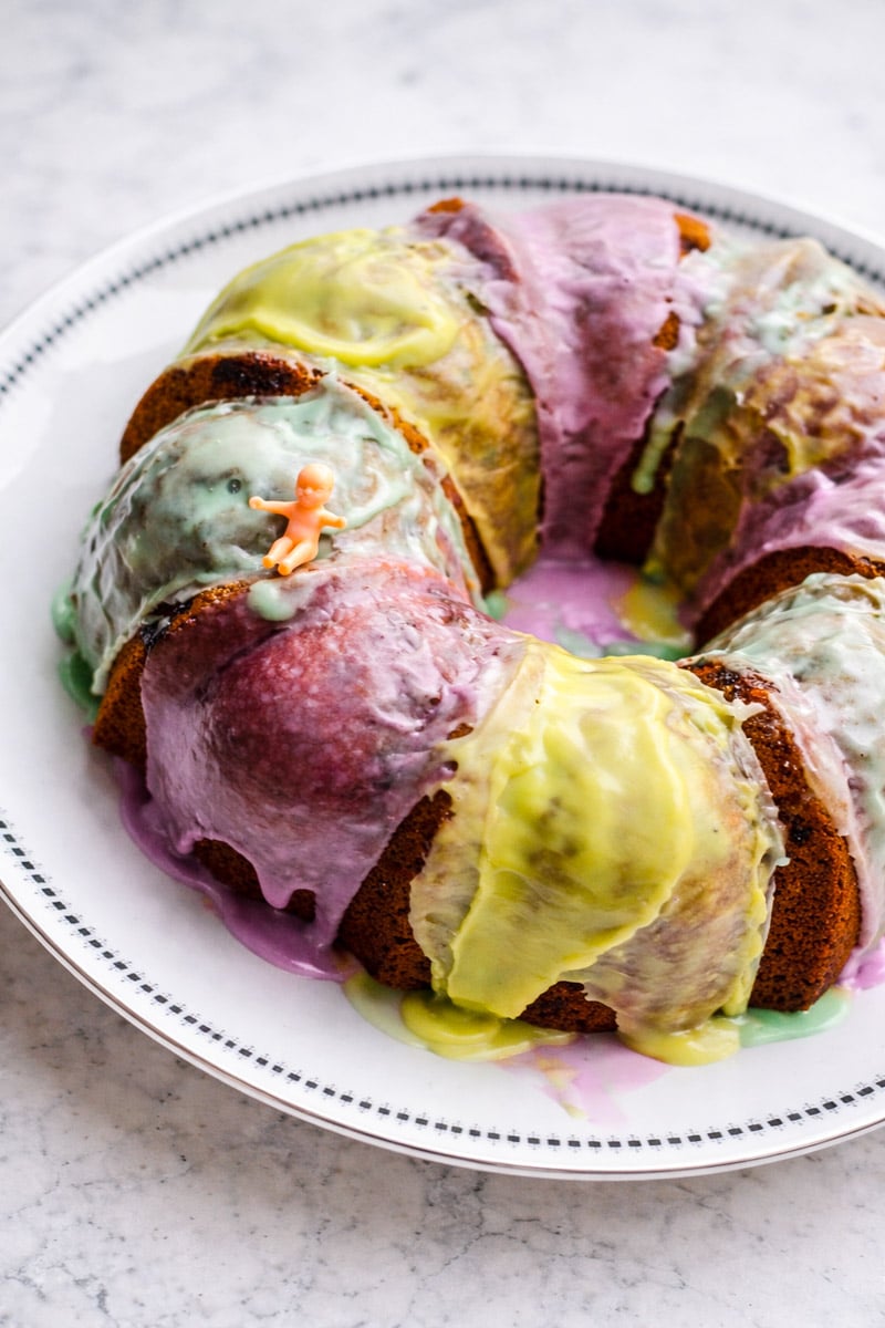 A King Cake with colorful purple, yellow, and green icing sits on a white plate. A small plastic baby figurine is placed on top of the cake.